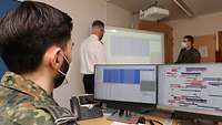 Ein Mann sitzt an bei Computer-Monitoren, im Hintergrund stehen zwei weitere Männer in Uniform.