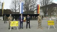 Zwei Soldaten und zwei Zivilisten stehen hinter Ortstafeln, die auf Dreibeine gestellt sind; im Hintergrund sitzen Zuschauer.