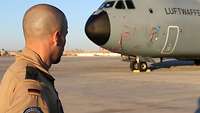 Im Vordergrund steht ein Soldat mit dem Rücken zum Bild. Im Hintergrund steht ein Transportflugzeug auf der Landebahn.
