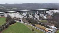 Eine lange, hohe Betonbrücke fällt in Staubwolken zusammen, ihre Pfeiler knicken ein. Dahinter steht eine zweite Brücke.