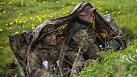 Zwei Soldaten mit Tarnschminke im Gesicht liegen unter einer tarnfarbenen Plane auf einer Wiese.