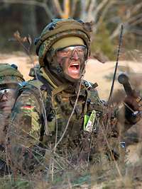 Zwei Soldaten hocken im Gras hintereinander, einer schreit ein Kommando.