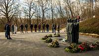 Eine Gruppe von Personen steht im Halbkreis um eine Skulptur, vor der Blumenkränze liegen.