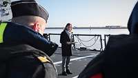 Eine Frau in Mantel und Schal steht auf dem Flugdeck eines Marineschiffs.