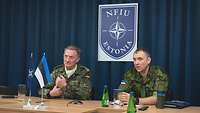 Zwei Soldaten sitzen an einem langen Tisch nebeneinander, vor ihnen Wimpel, hinter ihnen ein Wappen.