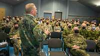 Zahlreiche Soldaten sitzen in einer Halle, vor ihnen steht ein General und spricht zu ihnen.