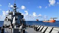 Eine Bohrplattform und ein Schiff im Hintergrund; davor das vordere Oberdeck eines grauen Kriegsschiffs.