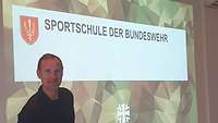 Oberstleutnant Gidion steht in sportlicher Kleidung vor der Leinwand mit dem Thema Sport für Soldaten mit Behinderung.