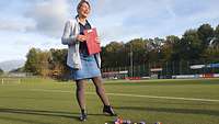 Eine Sportlehrerin steht in sportlicher Kleidung mit einem Klemmbrett auf dem Sportplatz.
