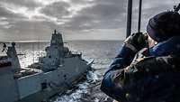 Ein graues Kriegsschiff in See; im Vordergrund ein Marinesoldat in blauer Arbeitsjacke mit einem Fernglas.