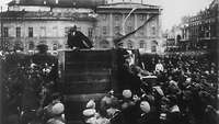 Schwarz-Weiß-Aufnahme: Lenin spricht von einer Tribüne zu vielen Menschen. Auf der Treppe der Tribüne stehen zwei Männer.