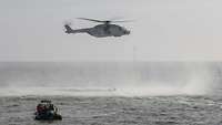 Ein grauer Hubschrauber schwebt in einer festen Position über dem Meer; auf der Wasseroberfläche sind Wirbel zu sehen.