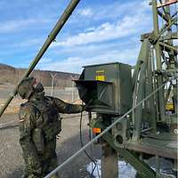 Im Vordergrund steht ein Soldat mit Helm und Nase-Mund-Bedeckung. Hinter ihm ist eine große militärische Antenne aufgebaut.