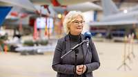 Ministerin Lambrecht steht in einer Halle und spricht in ein Mikrofon. Im Hintergrund stehen Kampfflugzeuge.