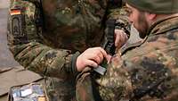 Ein Soldat legt eine schwarze Schlinge um den Oberarm eines anderen Soldaten.