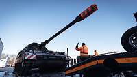 Eine Panzerhaubitze fährt auf einen LKW-Auflieger. Vor ihr steht ein Soldat in orangener Jacke und gibt Zeichen.