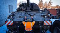 Eine Panzerhaubitze steht mittig auf einem Lastkraftwagen-Auflieger. Davor steht ein Soldat in Warnweste und hebt beide Arme.
