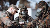 Zwei Soldaten in ABC-Schutzausstattung werden durch einen Soldaten in normaler Bekleidung angeleitet.