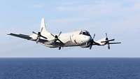 Ein graues Flugzeug mit vier Propellern fliegt über ein Meer.