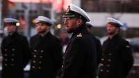 Ein Offizier der Marine in Uniform. Im Hintergrund stehen Soldaten in Reihe angetreten.