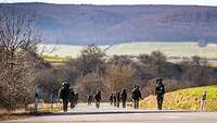 Soldaten marschieren in zwei Reihen auf einer Asphaltstraße durch hügliges Gelände in der Sonne.