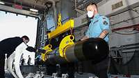 Drei Marinesoldaten in blauer Arbeitsuniform bewegen im Schiff einen grau-schwarzen Torpedo an einem gelben Kran.