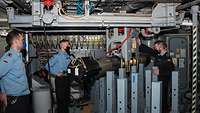 Drei Marinesoldaten in blauer Arbeitsuniform stehen in einem Raum; in der MItte hängt in einem Gestell von der Decke ein Torpedo