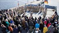 Auf dem Flugdeck eines Kriegsschiffs sind Soldaten angetreten; im Vordergrund eine zuschauende Menschenmenge.