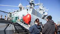 Eine Frau hält einen roten, herzförmigen Ballon in der Hand; im Hintergrund ein graues Kriegsschiff.