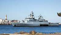 Ein graues Kriegsschiff auf Wasser; im Hintergrund Gebäude einer Hafenstadt.