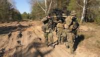 Drei Soldaten transportieren auf ihren Schultern eine Granatmaschinenwaffe durch bewaldetes Gelände.