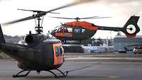 Ein Hubschrauber steht auf einem Flugplatz, dahinter schwebt ein weiterer ein.