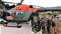 Mehrere Soldaten laden einen Verletzten auf einer Trage in einen orange-grünen Hubschrauber.