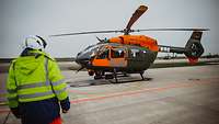 Ein Soldat in signalgrüner Jacke steht auf einem Flugplatz vor einem orange-grünen Hubschrauber.