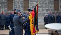 Ein Soldat übergibt einem weiteren Soldaten eine Flagge. Im Hintergrund andere Soldaten und Zuschauende.