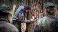 Ein Soldat zeigt den anderen Soldatinnen und Soldaten wie man die Rinde vom Baum abschlägt.