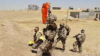 Ein Soldat hält in der Wüste ein orangefarbenes Identifikationstuch in die Luft, weitere Soldaten sichern im Hintergrund