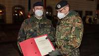 Abends auf einem Marktplatz: Ein Soldat überreicht einem zweiten eine Urkunde.