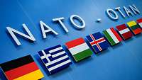 Unter dem Schriftzug NATO – OTAN sind mehrere Flaggen von Mitgliedstaaten zu sehen.