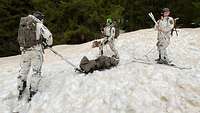 Drei Soldaten in Schneetarn mit Skiern ziehen eine Trage mit einem Verletzten am verschneiten Berg.