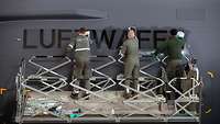 Three soldiers stand on a lift truck and clean the lettering ‘LUFTWAFFE’ on the fuselage of the C-130J with rags.