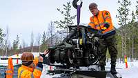 Zwei Soldaten mit orangefarbenen Jacken heben mit einem Kran einen Motor aus einem Fahrzeug.