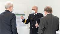 Zwei Männer und ein Soldat stehen vor einem Whiteboard und diskutieren