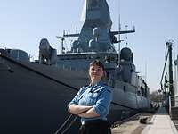 Eine Soldatin steht mit verschränkten Armen vor einem grauen Schiff.