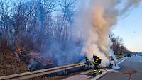 Feuerwehrleute löschen ein brennendes Auto, das auf dem Standstreifen einer Autobahn steht.