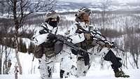 Zwei bewaffnete Soldaten in Schneetarnuniform abgekniet im Schnee. Im Hintergrund eine weitläufige Winterlandschaft und Berge.