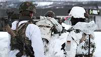 Zwei Soldaten in Schneetarnuniform blicken auf einen Soldaten der hinter einem Schweren Maschinengewehr liegt.