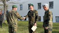 Zwei Soldaten stehen vor einem hellen Gebäude, einer reicht einem dritten Soldaten die Hand.