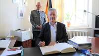 Ein Mann sitzt am Schreibtisch und schreibt in ein Buch. Ein weiterer Mann steht dahinter
