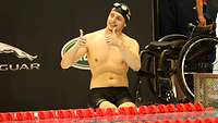 Ein deutscher Schwimmsportler sitzt am Schwimmbeckenrand und hält die Daumen hoch. Neben ihm steht ein Rollstuhl.